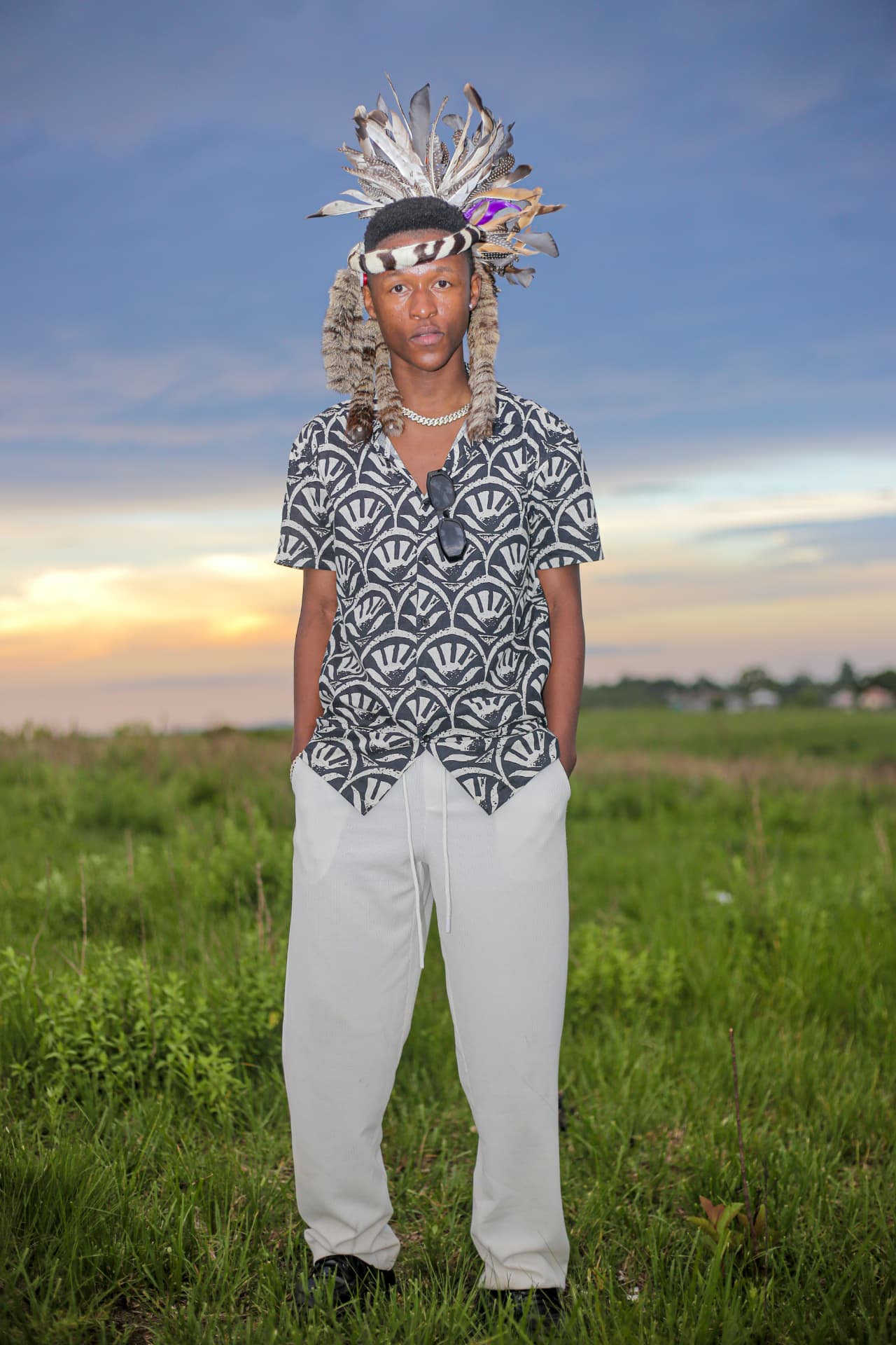 Portrait of a young man in a feathered Zulu headdress standing in open grassland at sunset
