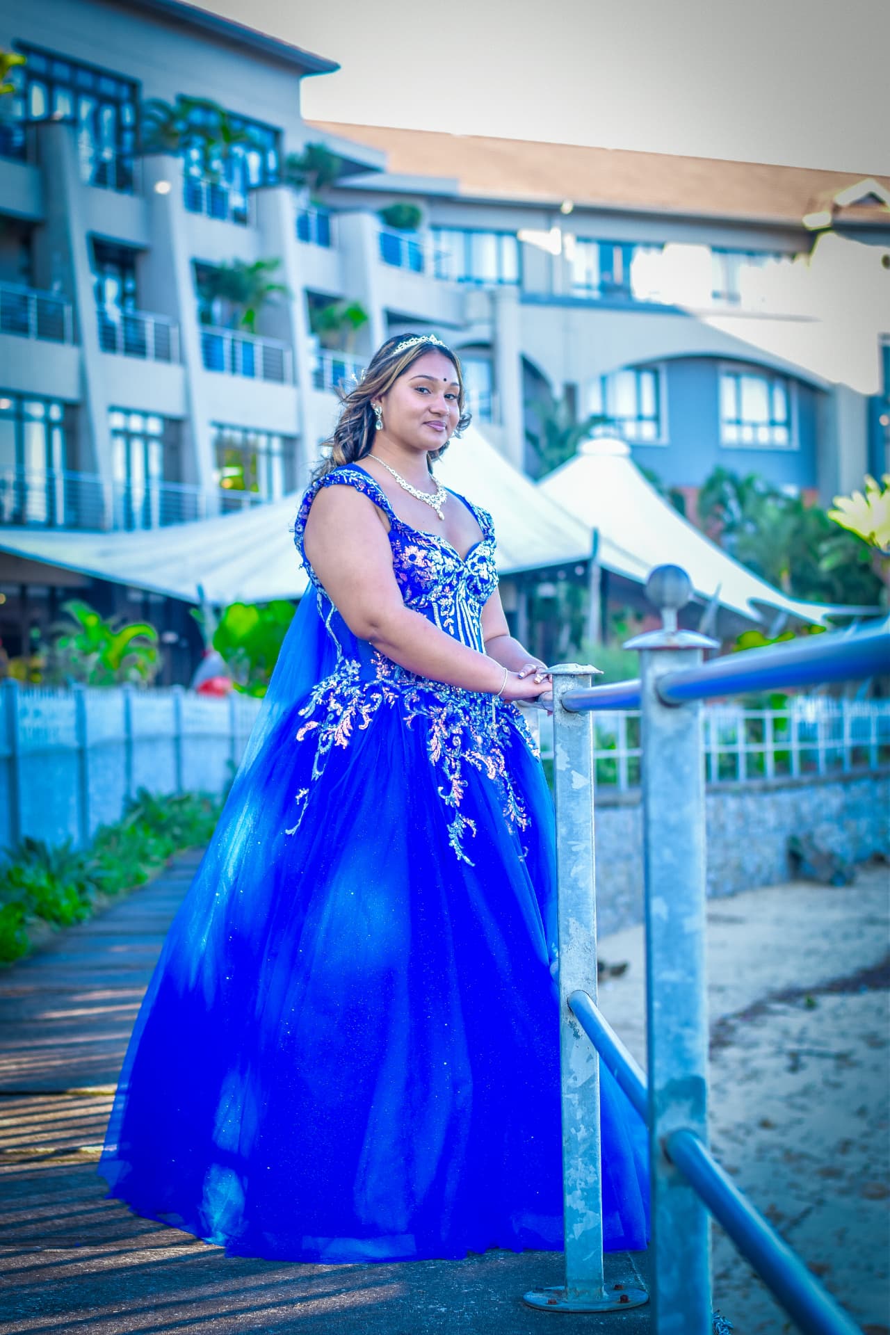 Debutante in a sapphire beaded gown resting on a boardwalk rail near the shore