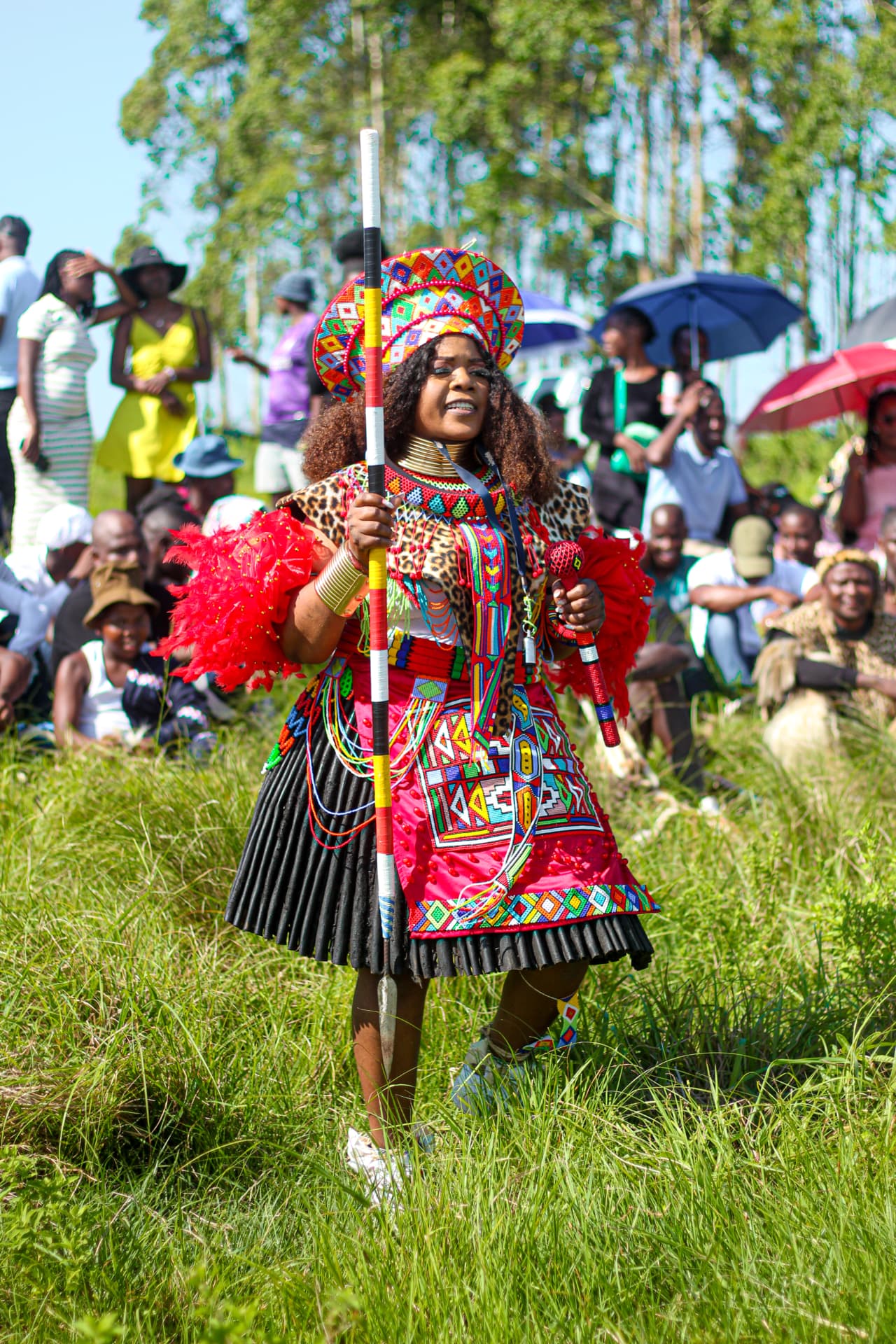 Celebrant in full Zulu regalia holding a ceremonial staff before a seated crowd