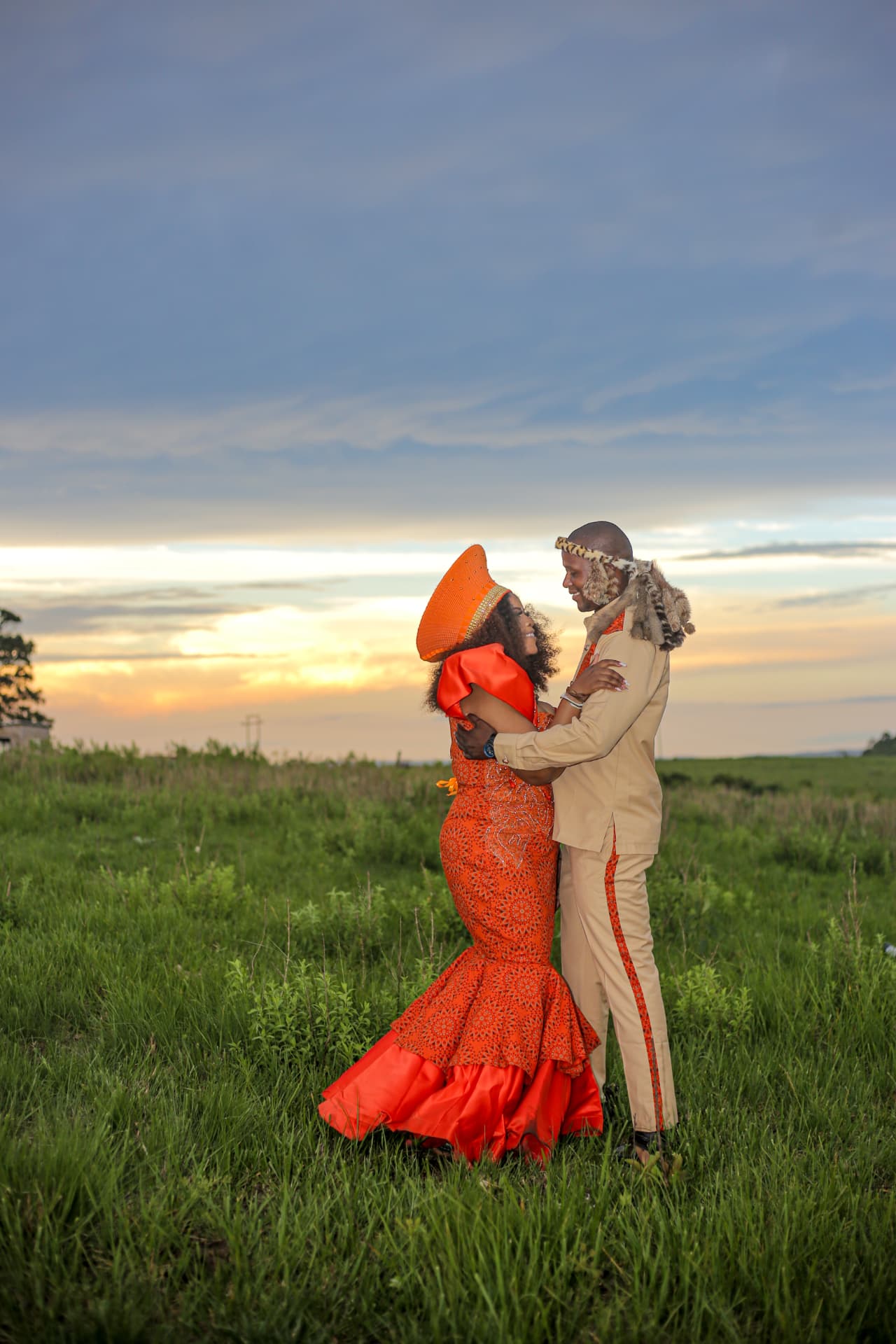 Bride and groom in traditional attire embracing in open grassland at dusk